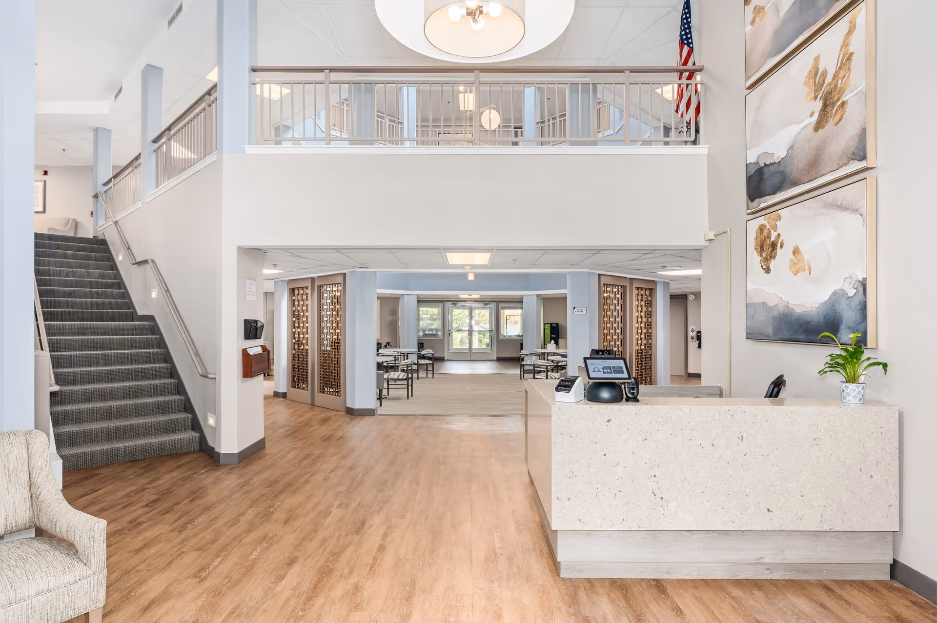 Bright senior living facility lobby with a reception desk, staircase, seating and a view into a dining/activity area.