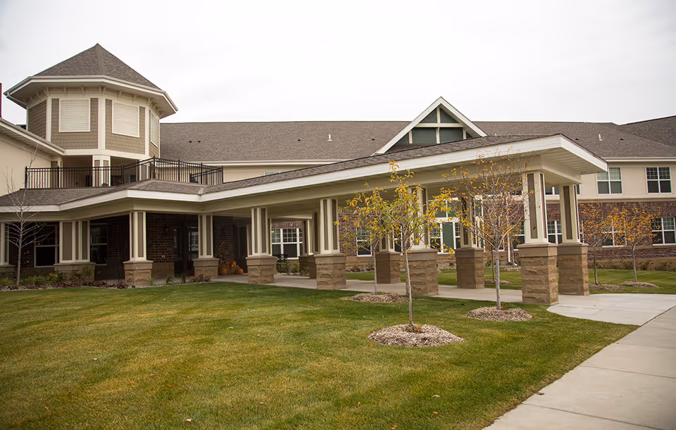 Front exterior of a senior living building with a covered entrance, lawn, and small trees.