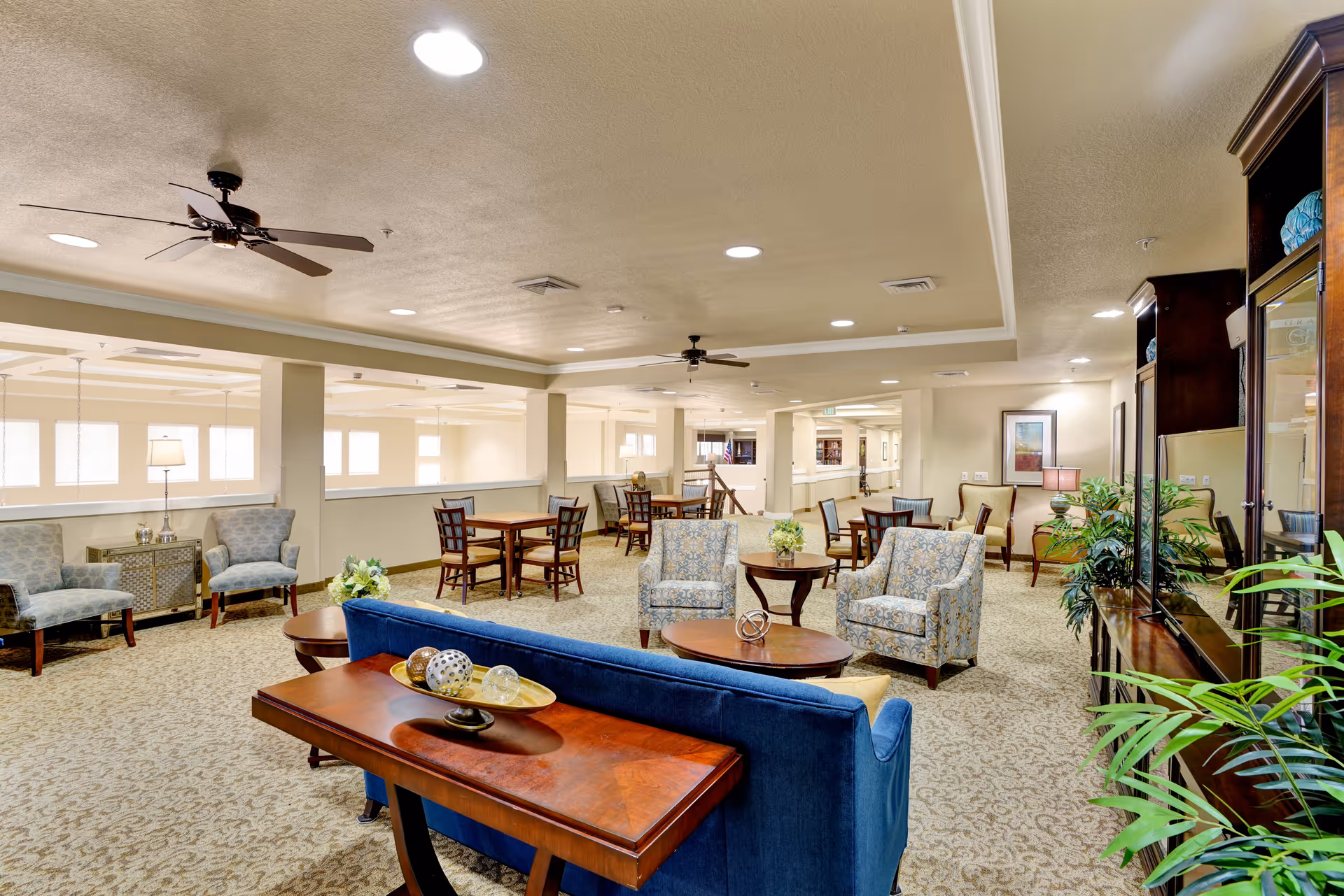 A spacious and well-lit senior living facility common area with multiple seating arrangements including a blue sofa, patterned armchairs, wooden tables, and chairs. The room features ceiling fans, recessed lighting, carpeted floors, decorative plants, and framed artwork on the walls.