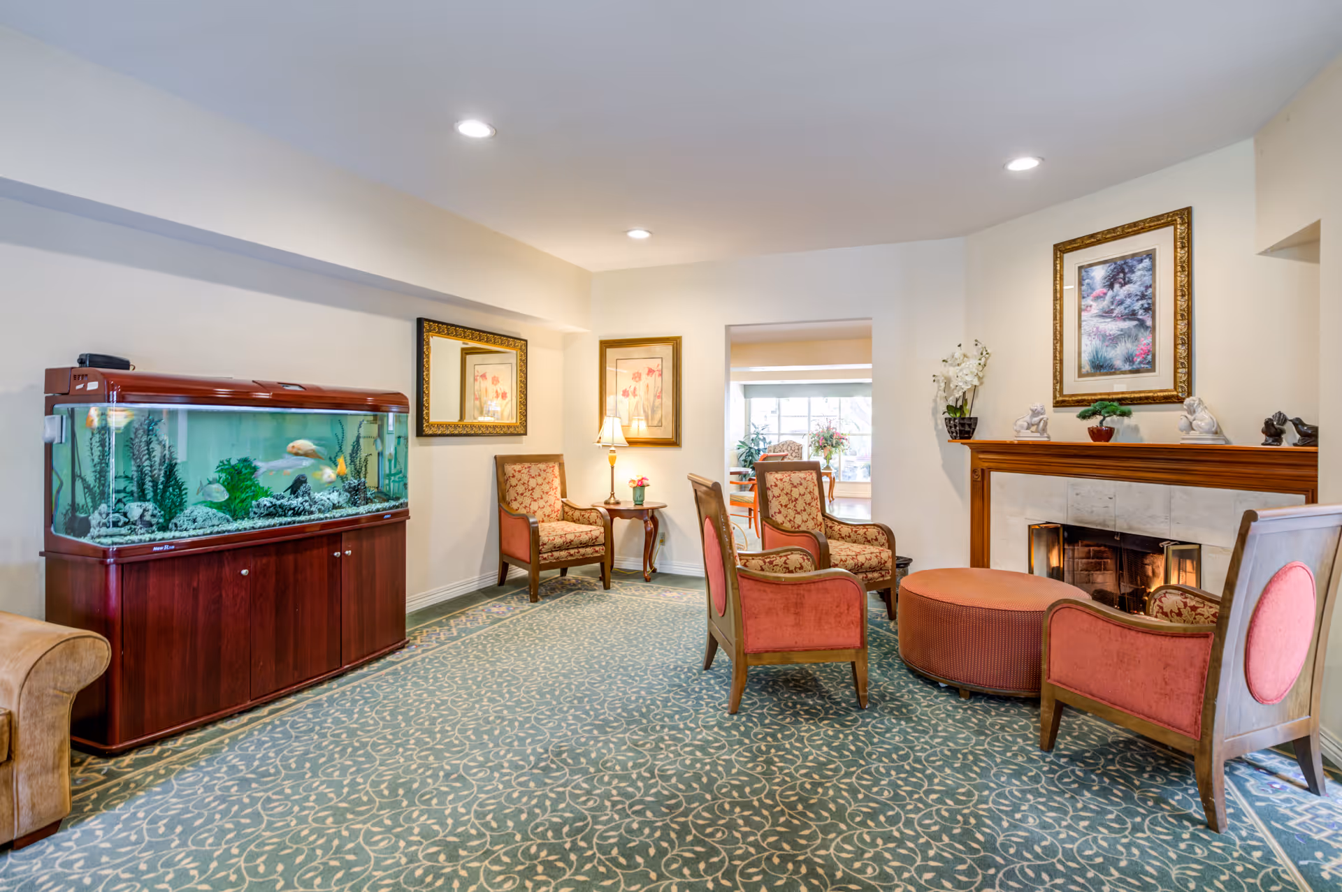 A cozy living room area with patterned carpet, four upholstered chairs arranged around a round ottoman, a fireplace with decorative items on the mantel, framed artwork on the walls, a large fish tank on a wooden cabinet, and a small side table with a lamp and flowers.
