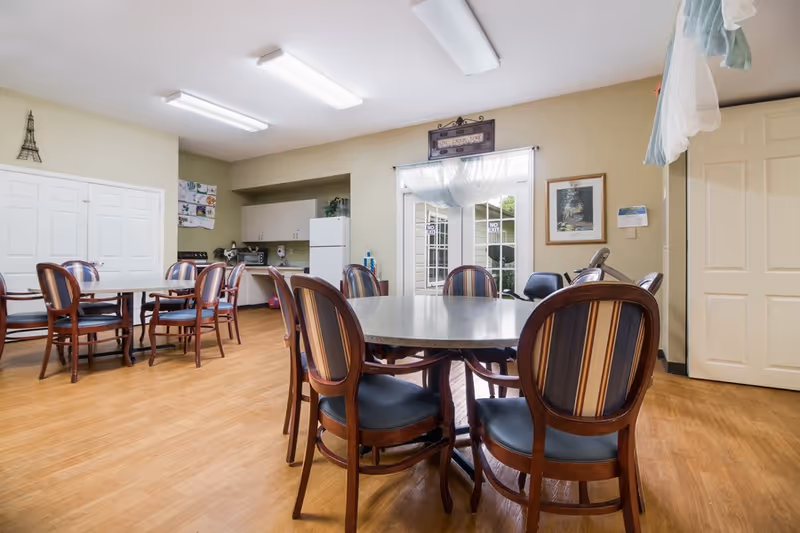 Bright communal dining/activity room with round tables and striped-back chairs, a small kitchenette, and double doors to the outside.