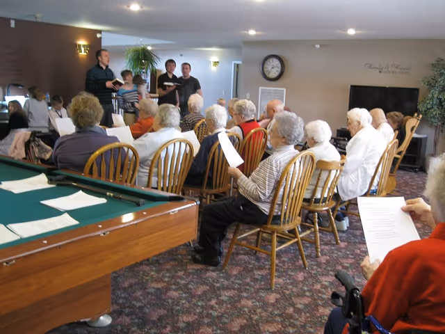 A group of elderly residents sit in chairs in a common room holding papers while a few younger people stand at the front, with a pool table visible in the foreground.
