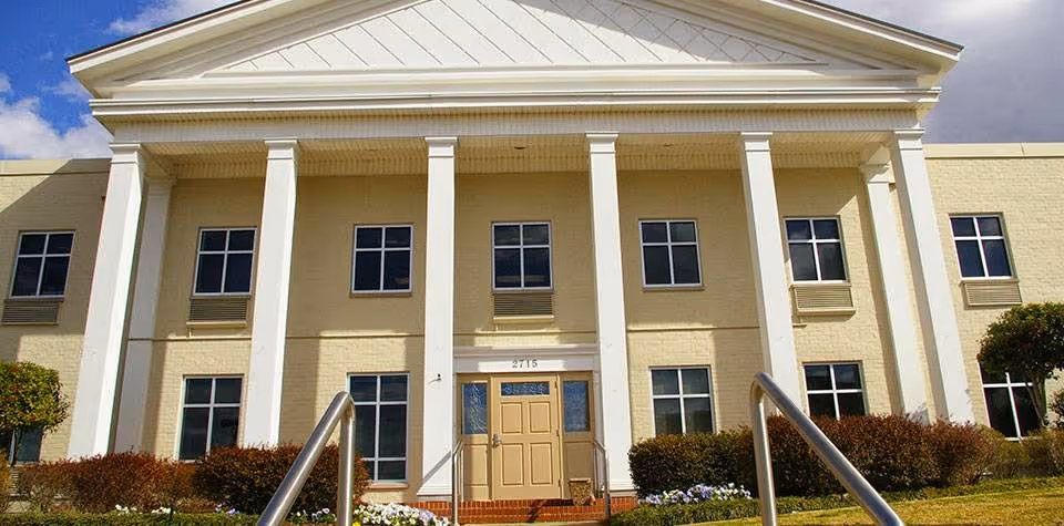 Front exterior view of a two-story building with beige brick walls, white columns, and a triangular pediment above the entrance. The building has multiple windows and a central door with the number 2715 above it. There are bushes and flowers in front of the building and a handrail leading up to the entrance.