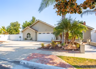 Single-story residential building with a two-car garage, surrounded by palm trees and other greenery, under a clear blue sky.