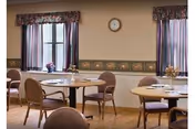 A dining room with round tables and chairs arranged neatly. Each table has a small centerpiece, and the room has two windows with floral curtains. A clock is mounted on the wall between the windows.