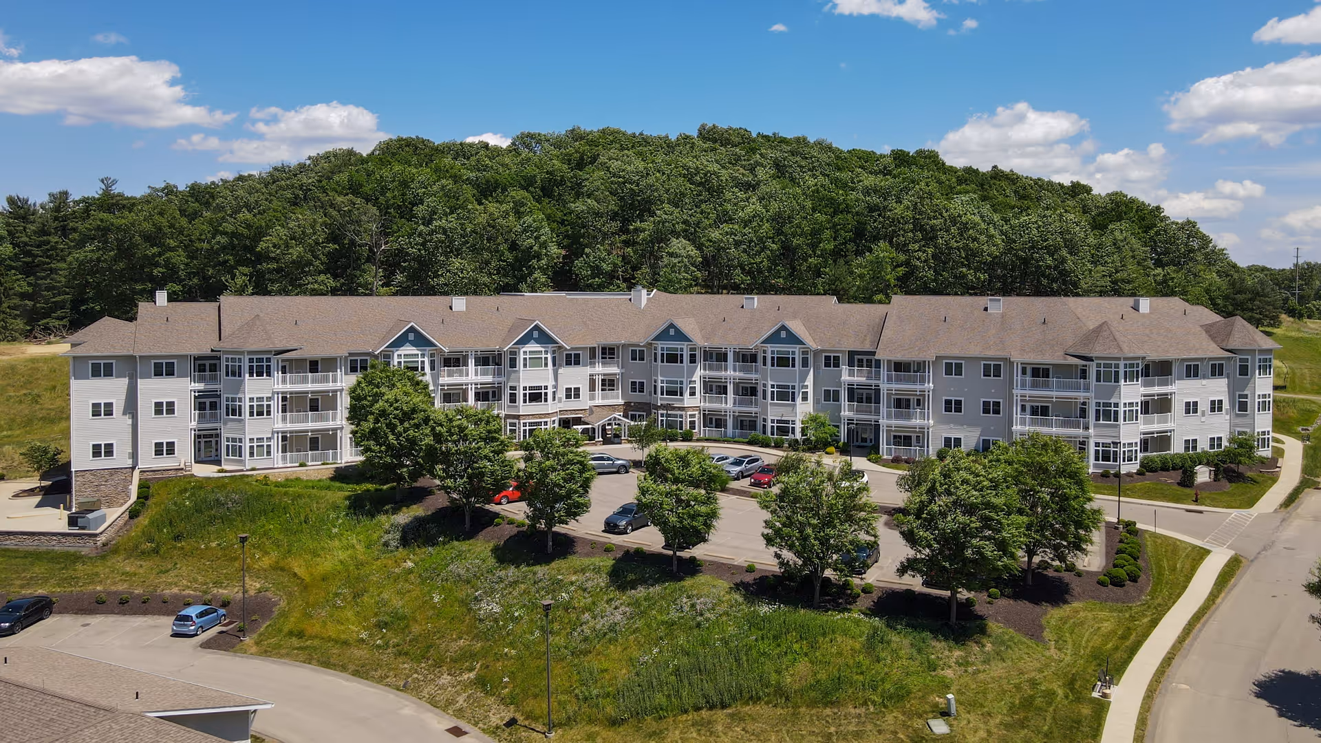 A large three-story senior living facility building with multiple balconies, surrounded by green trees and grassy areas under a partly cloudy blue sky. There is a parking lot with several cars in front of the building and a wooded hill in the background.