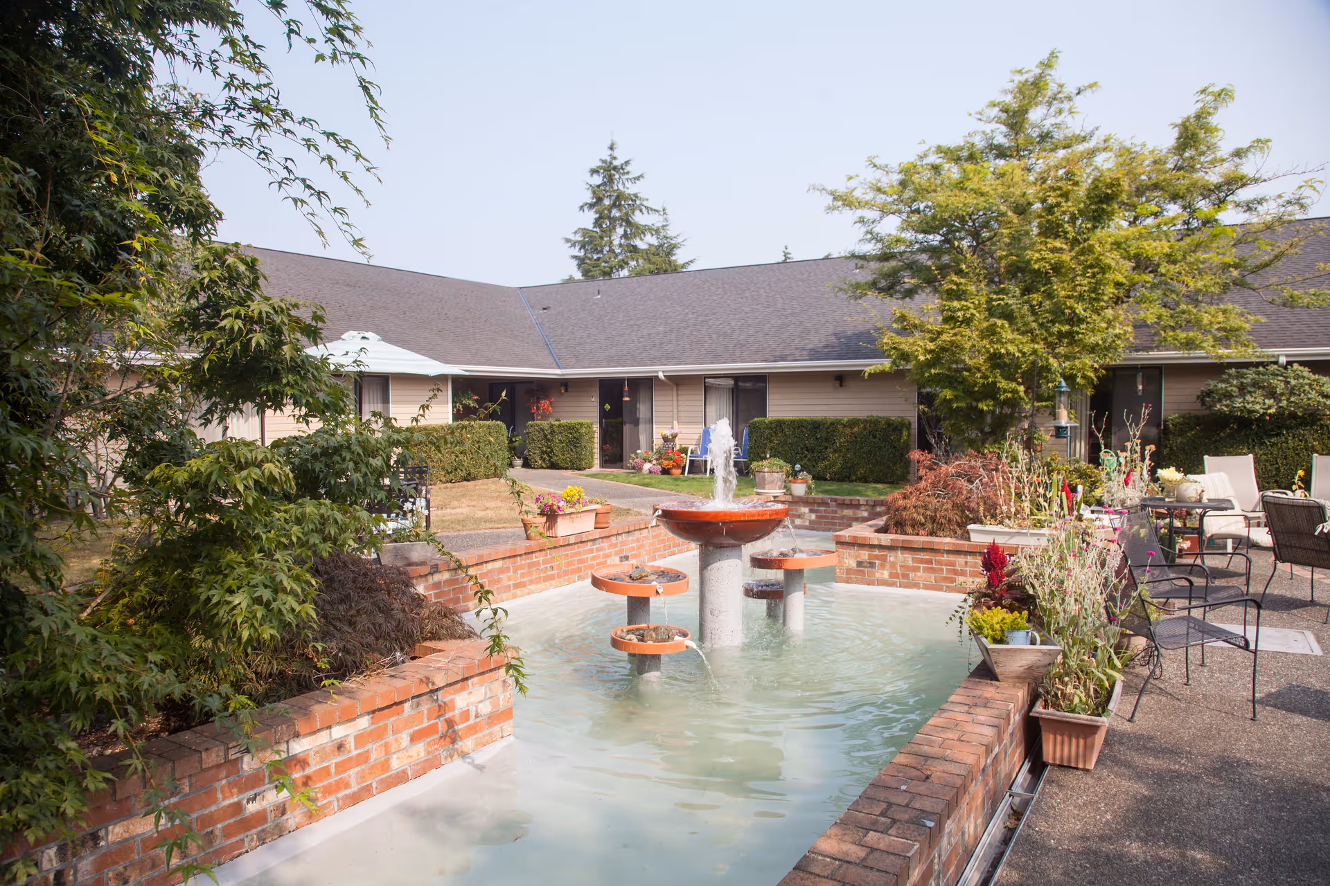 Outdoor courtyard area at Cap Sante Court Retirement Community featuring a brick water fountain with multiple tiers, surrounded by potted plants, shrubs, and patio chairs. The building with beige siding and dark roof is visible in the background under a clear sky.