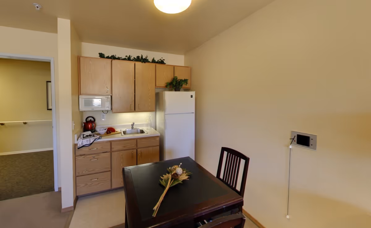 Small kitchenette with wooden cabinets, a sink, microwave and refrigerator next to a dining table and chairs.