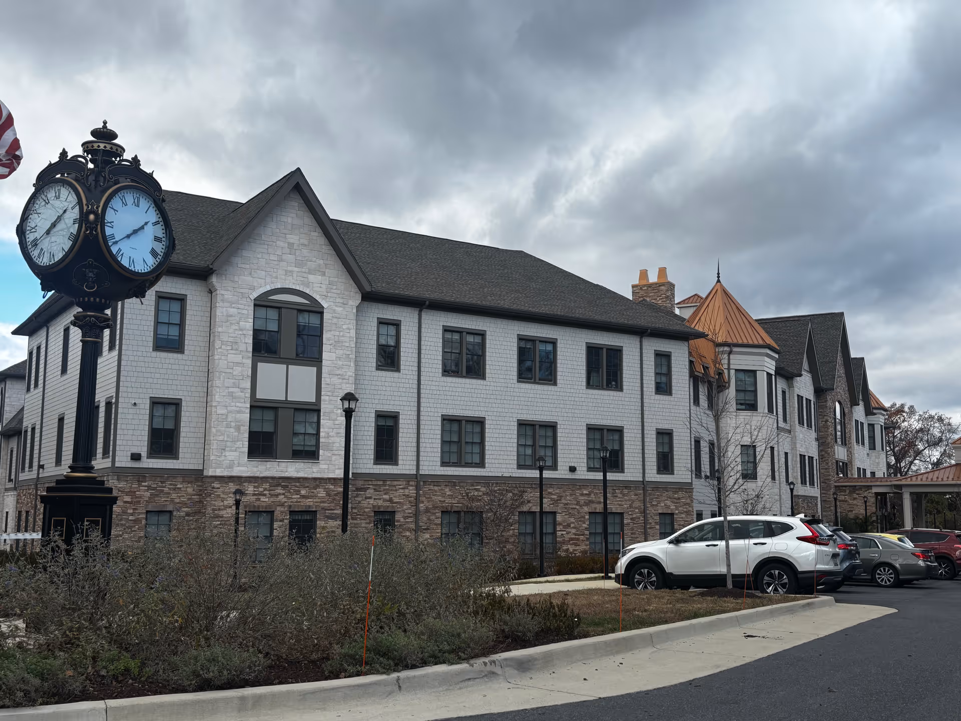 Exterior view of a multi-story senior living facility building with a mix of stone and shingle siding, multiple windows, and a peaked roof. There is a vintage-style street clock on a black post near the building, an American flag partially visible, several parked cars, and a cloudy sky overhead.