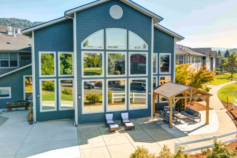 Exterior view of a modern building with large reflective windows and a small outdoor seating area under a wooden pergola. The building is surrounded by a paved walkway and landscaped greenery with trees and bushes.