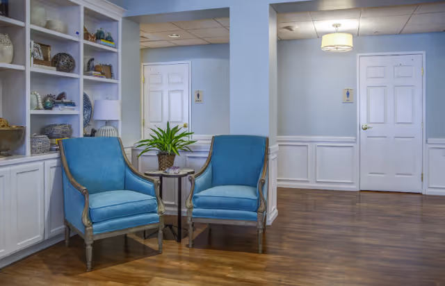 A cozy interior seating area with two blue upholstered armchairs and a small round table with a potted plant between them. The space has light blue walls, white wainscoting, wood flooring, and built-in white shelves with decorative items. Two white doors with restroom signs are visible in the background.