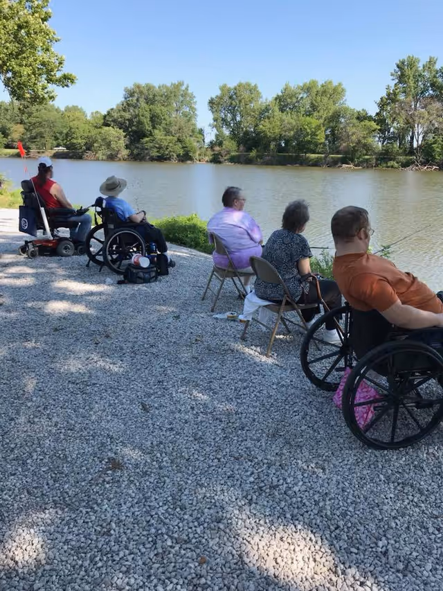Five people, some in wheelchairs and others seated on folding chairs, are fishing by a calm river on a sunny day with trees in the background.