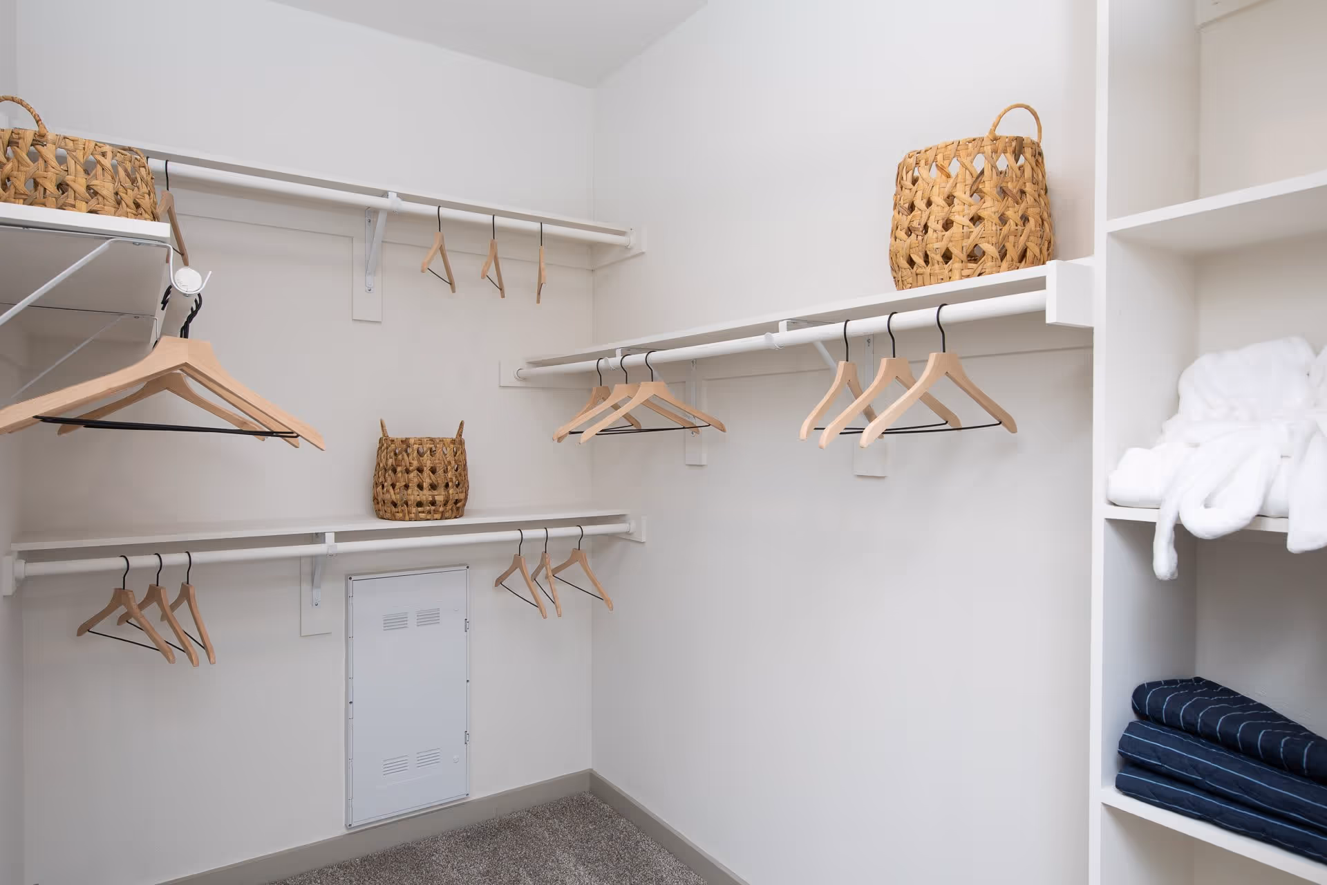 A spacious walk-in closet with white walls and multiple white shelves and hanging rods. Several wooden hangers are hanging on the rods, and two woven baskets are placed on the shelves. On the right side, folded white robes and dark blue blankets are neatly stacked on open shelving.