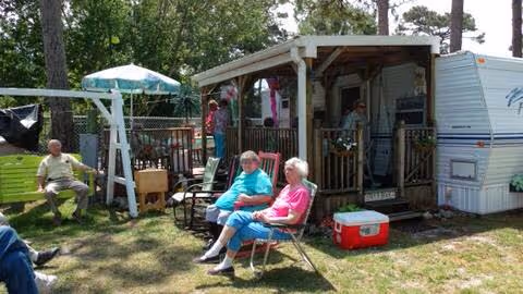 Three elderly people sitting and standing outside near a small wooden porch attached to a trailer. Two women are seated on lawn chairs in front of the porch, while a man is sitting on a swing to the left. There is a red cooler on the grass and a green umbrella providing shade in the background. Trees and grass surround the area.