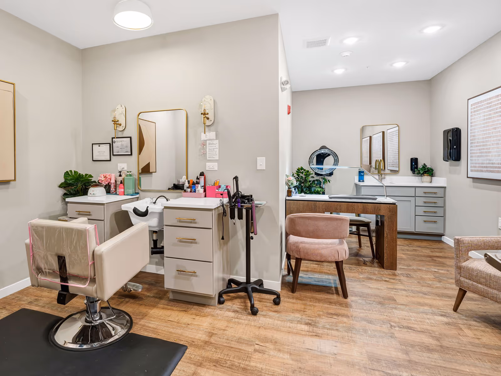 A well-lit salon area in a senior living facility featuring a styling chair with a protective cover, a sink for hair washing, a vanity with a mirror, and various hair care products. The room has wood flooring, neutral-colored walls, and additional seating with a pink cushioned chair and a small table with a lamp and plants. There are framed certificates and artwork on the walls.