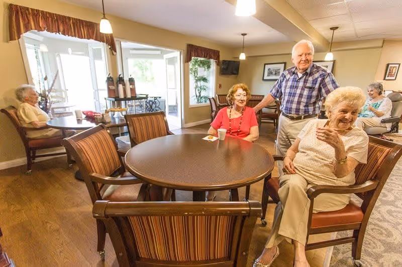 A group of elderly people sitting and standing around tables in a well-lit common area with large windows and wooden floors at Creekside Oaks Retirement Community.