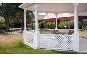 White gazebo with lattice railing in a grassy outdoor area near a red brick building surrounded by trees and shrubs.