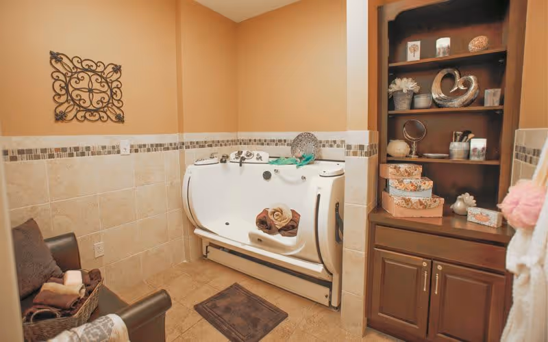 A bathroom with beige tiled walls and floor, featuring a white walk-in bathtub with rolled towels inside. To the right, there is a wooden cabinet with shelves holding decorative items and storage boxes. On the left, a brown leather chair with a basket of towels is visible. The walls are painted a warm beige color and decorated with a metal wall art piece.