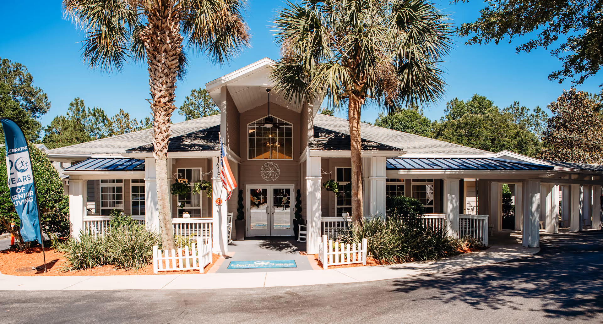 Front exterior view of The Gables of Gainesville senior living facility with palm trees, a small white picket fence, and an American flag near the entrance under a clear blue sky.