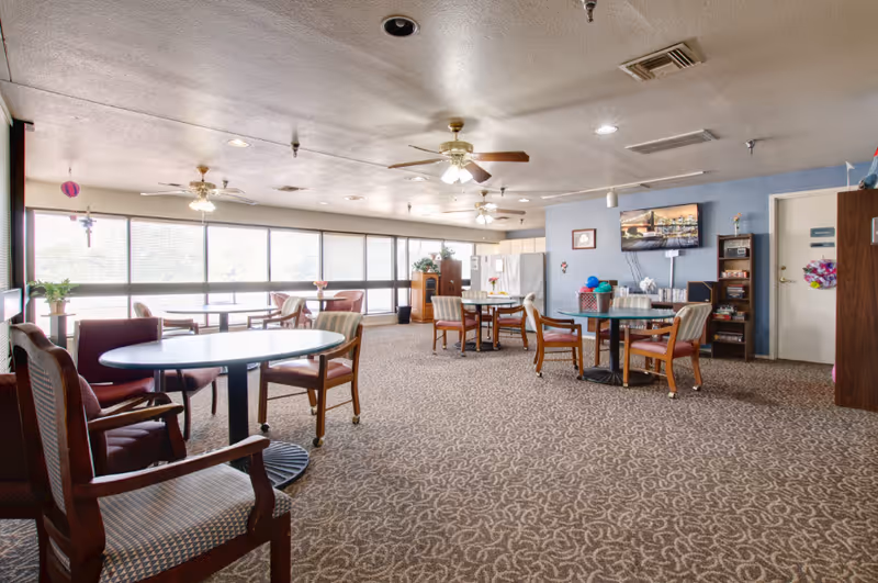 A spacious common area in an independent living facility with several round tables and chairs arranged on a patterned carpet. Large windows line one side of the room, allowing natural light to fill the space. Ceiling fans and recessed lighting are visible on the ceiling. A television is mounted on a blue wall, with shelves and decorations nearby.