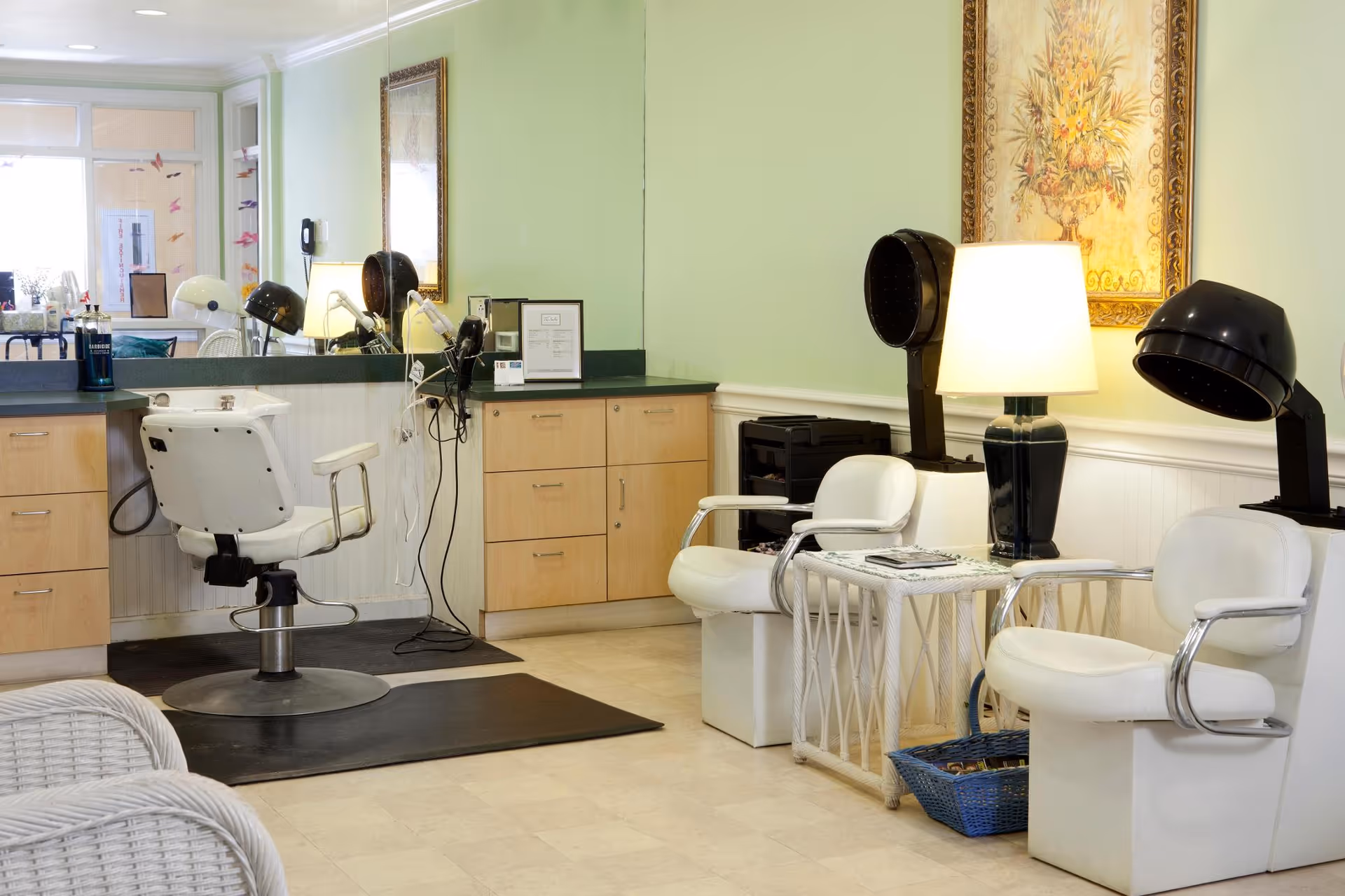 Interior view of a salon area in a senior living facility with a white salon chair in front of a mirror and counter, two white chairs with black hair dryers, a small table with a lamp and magazines, and a framed floral painting on the wall.