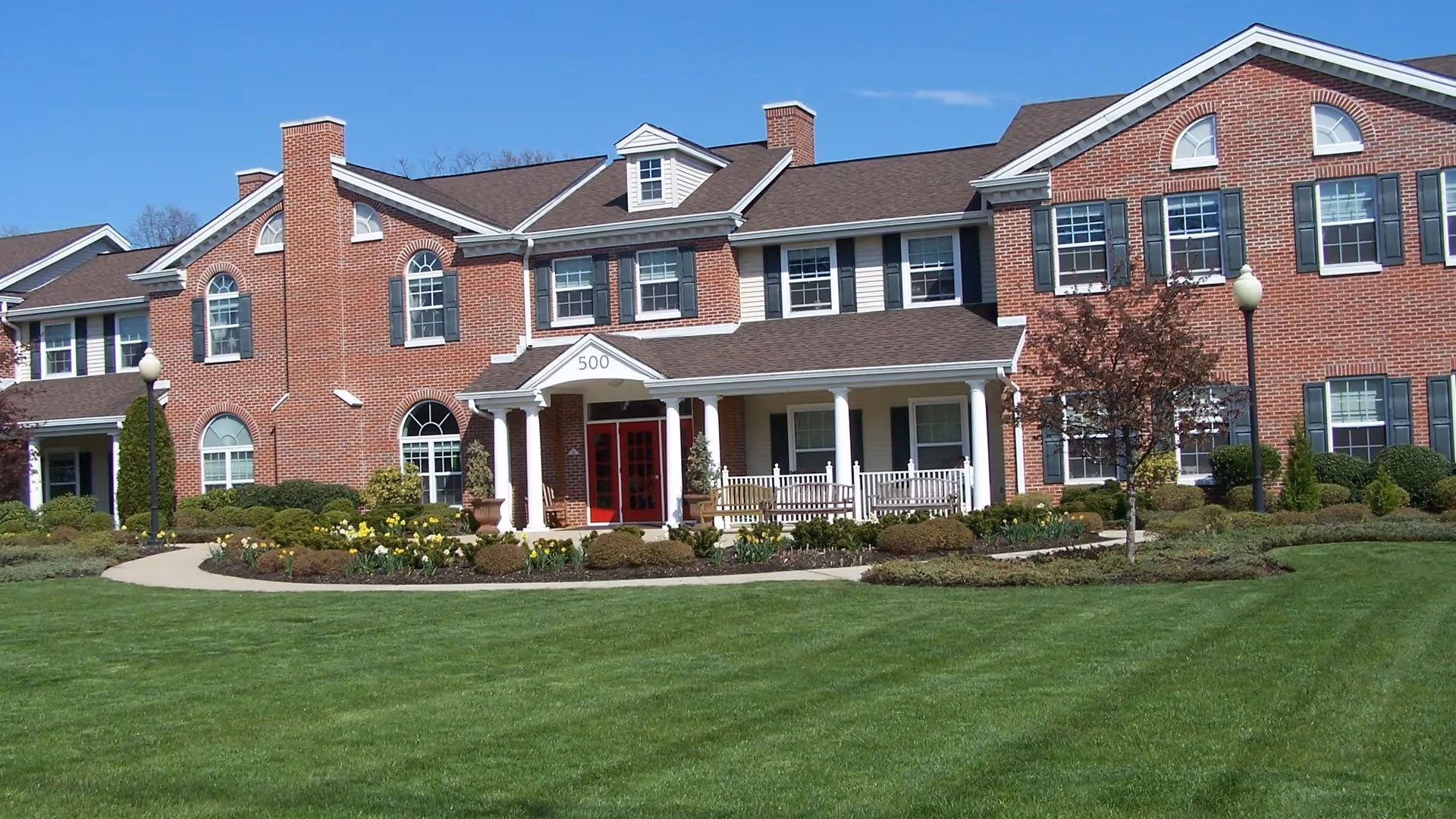Front exterior view of a large two-story brick building with white columns and a covered porch. The building has multiple windows with black shutters, a red double-door entrance marked with the number 500, and well-maintained landscaping with green grass, bushes, and flowers under a clear blue sky.
