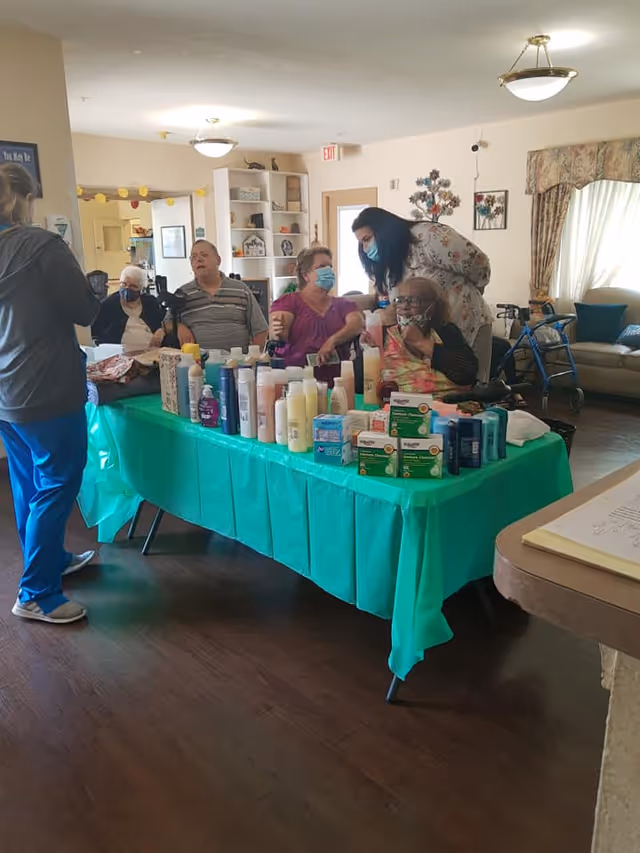 A group of elderly people sitting and standing around a table covered with a green tablecloth displaying various personal care products such as lotions, shampoos, and boxes of items. One caregiver wearing a mask is interacting with the seated individuals in a well-lit room with wooden floors, a couch, and decorative wall art.