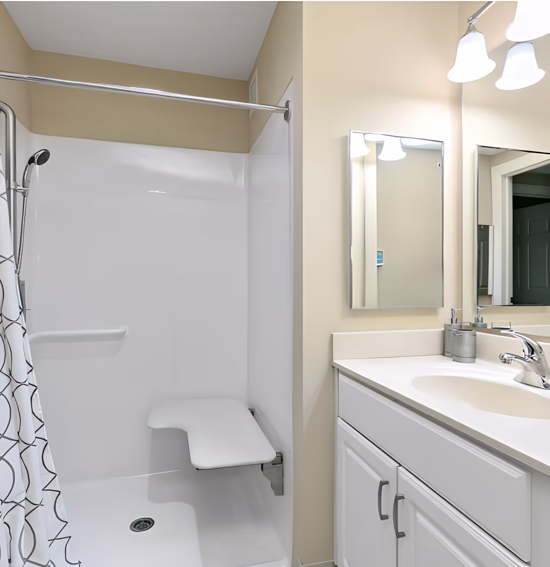 A clean and modern bathroom featuring a white shower area with a built-in foldable seat and a handheld showerhead. The shower has a curtain with a black and white geometric pattern. Adjacent to the shower is a white vanity with a sink, a chrome faucet, a soap dispenser, and a mirror above it. The walls are painted in a light beige color, and there are three light fixtures above the mirror providing bright illumination.