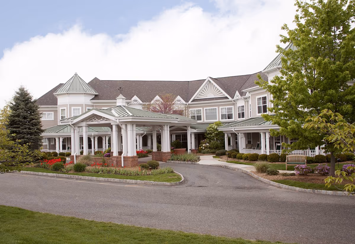 Front entrance of a large two-story senior living building with a covered porte-cochere, landscaped flowerbeds and a circular driveway.