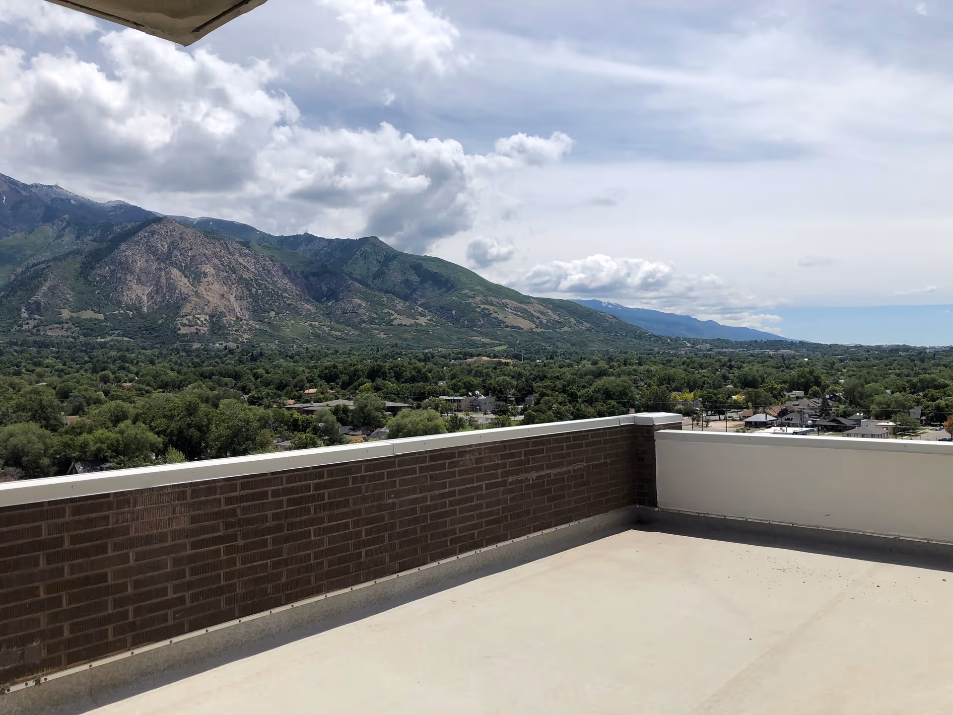 Rooftop terrace with a low brick railing overlooking a tree-filled valley and mountains under a cloudy sky.