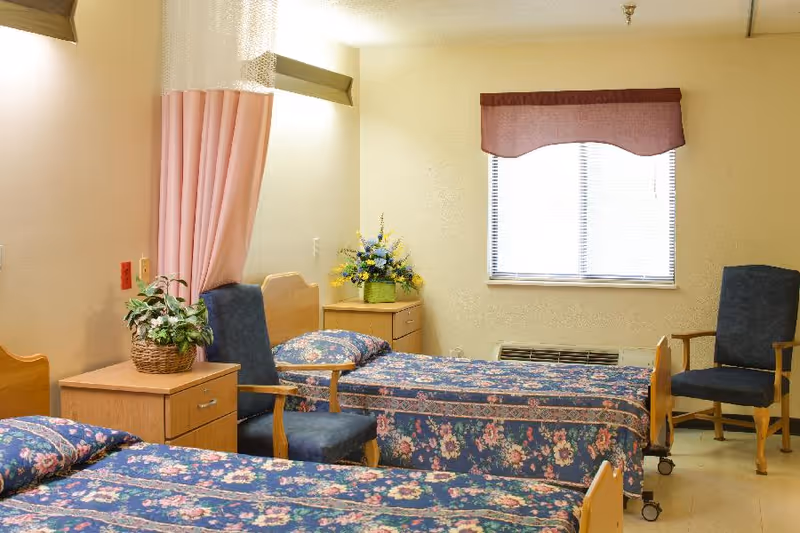 Shared resident bedroom with two floral-covered beds, chairs, a nightstand, and a window.