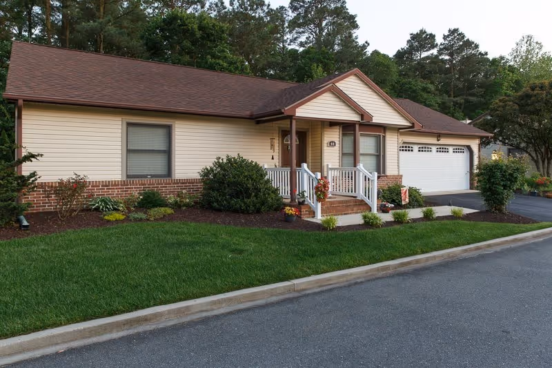 Single-story residential building with beige siding and brick accents, a brown shingled roof, a small front porch with white railings, and a two-car garage. The front yard has neatly trimmed green grass, bushes, and small plants. Trees are visible in the background.