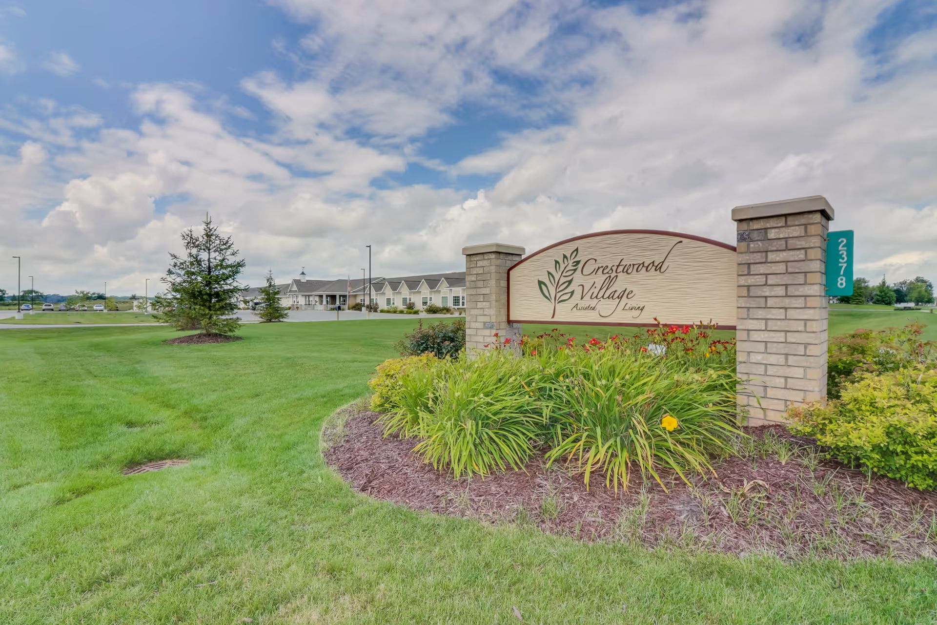Outdoor view of Crestwood Village Assisted Living & Memory Care facility sign surrounded by green grass and plants, with the building visible in the background under a partly cloudy sky.