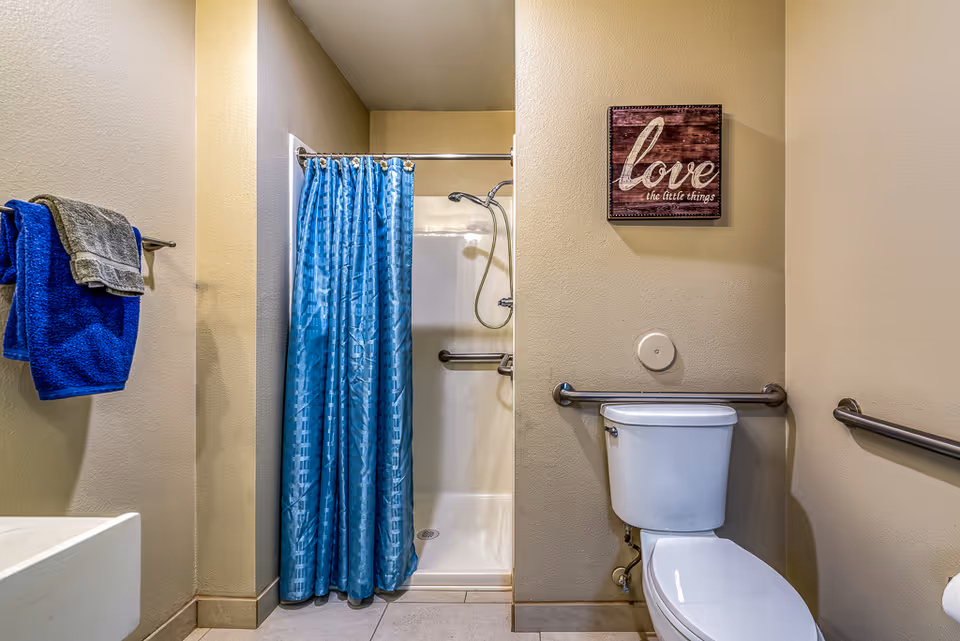 A bathroom with beige walls featuring a white toilet with safety grab bars on both sides, a shower with a blue curtain, and a towel rack holding blue and gray towels. A decorative sign on the wall reads 'love the little things.'