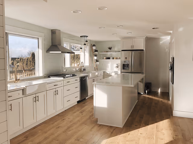 Bright modern kitchen with white cabinets, a large island with a white countertop, stainless steel refrigerator, dishwasher, and stove. The kitchen has wooden flooring, two large windows letting in natural light, and open shelves with plants and kitchenware.