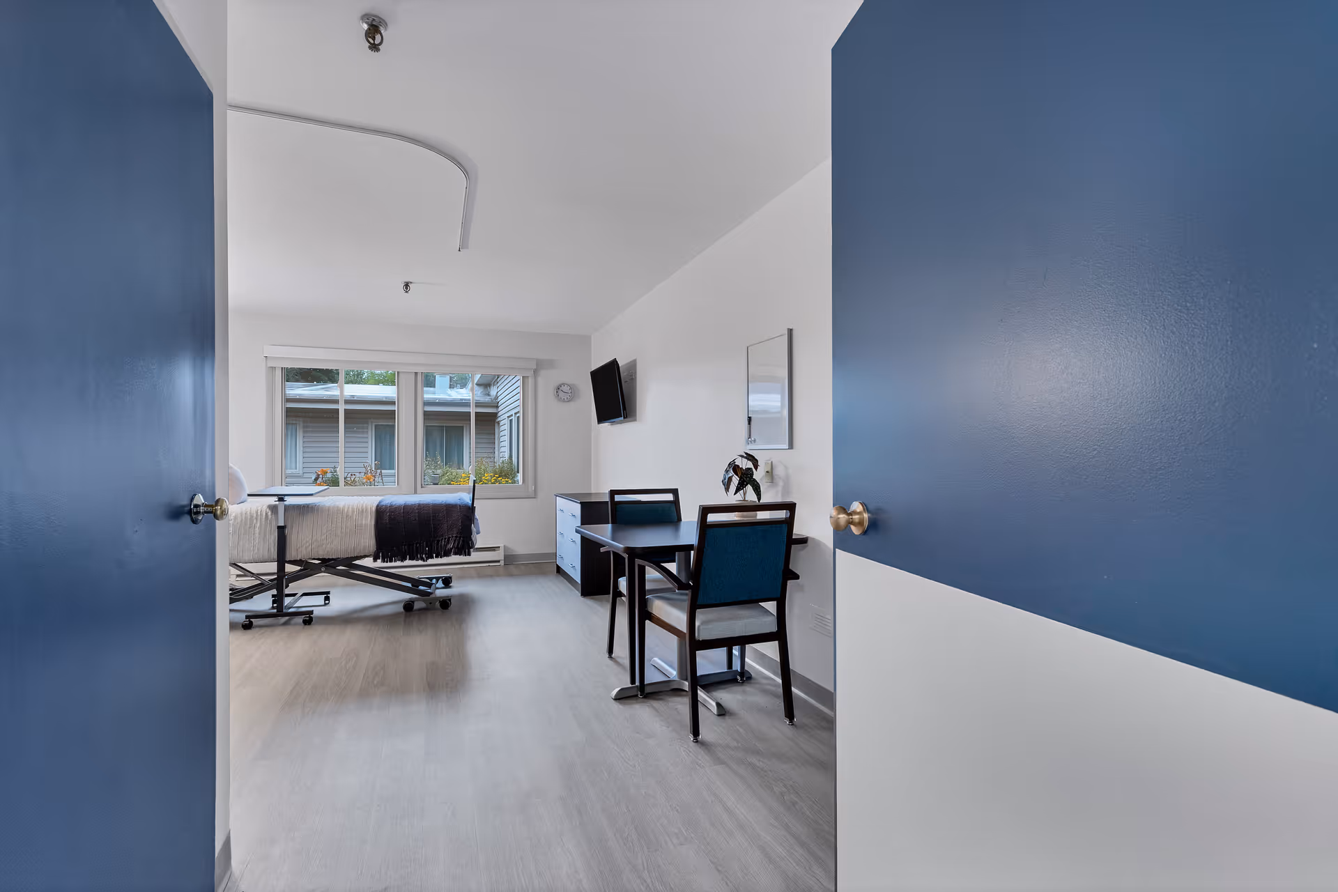 View through partially open double doors into a bright room with a hospital-style bed near a large window, a small table with two chairs, a wall-mounted TV, and a dresser. The room has light-colored flooring and white walls with blue doors.