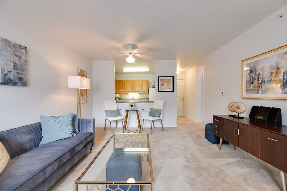 A bright and clean living room area in Oakhaven Manor featuring a blue sofa with cushions, a glass coffee table with a geometric gold frame, a wooden sideboard with decorative items and a stereo, two white chairs with green cushions around a small round table, and a view into a kitchen with wooden cabinets and white appliances. The walls are white with framed artwork, and the floor is carpeted.