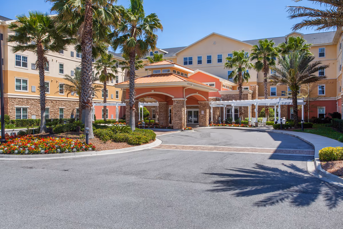 Front exterior view of a senior living facility named Camellia At Deerwood with a circular driveway, palm trees, colorful flower beds, and a covered entrance with stone pillars under a clear blue sky.