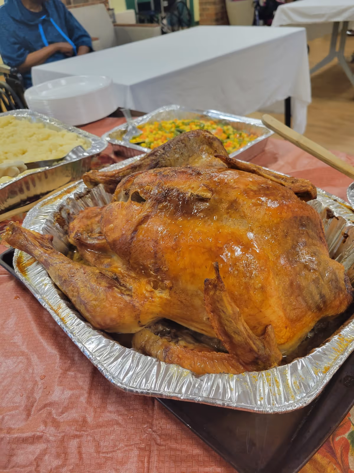 A golden roasted turkey in an aluminum tray on a table with mashed potatoes and mixed vegetables and a white-clothed table in the background.