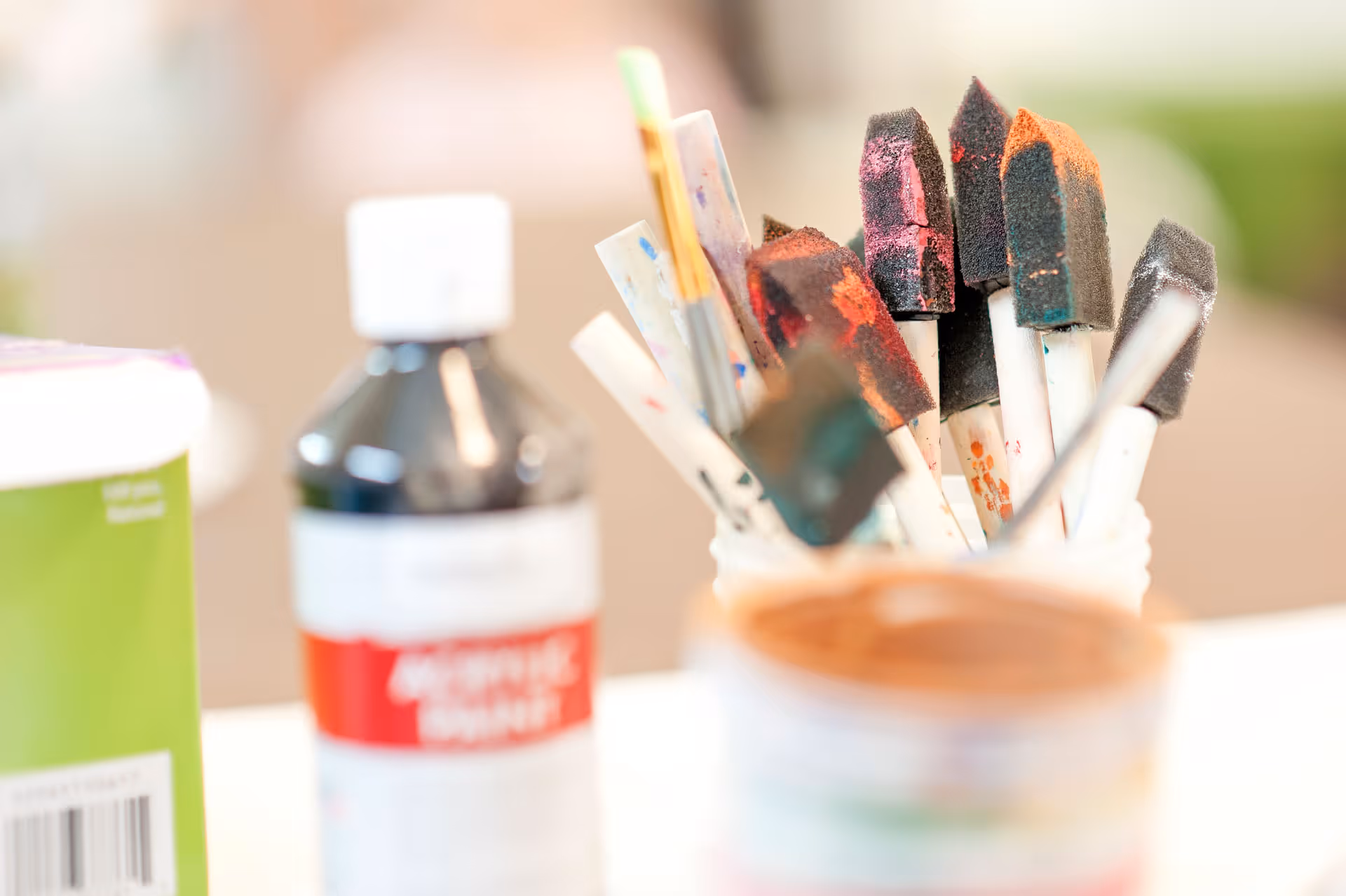 Foam paintbrushes in a container with blurred paint bottles on a table.
