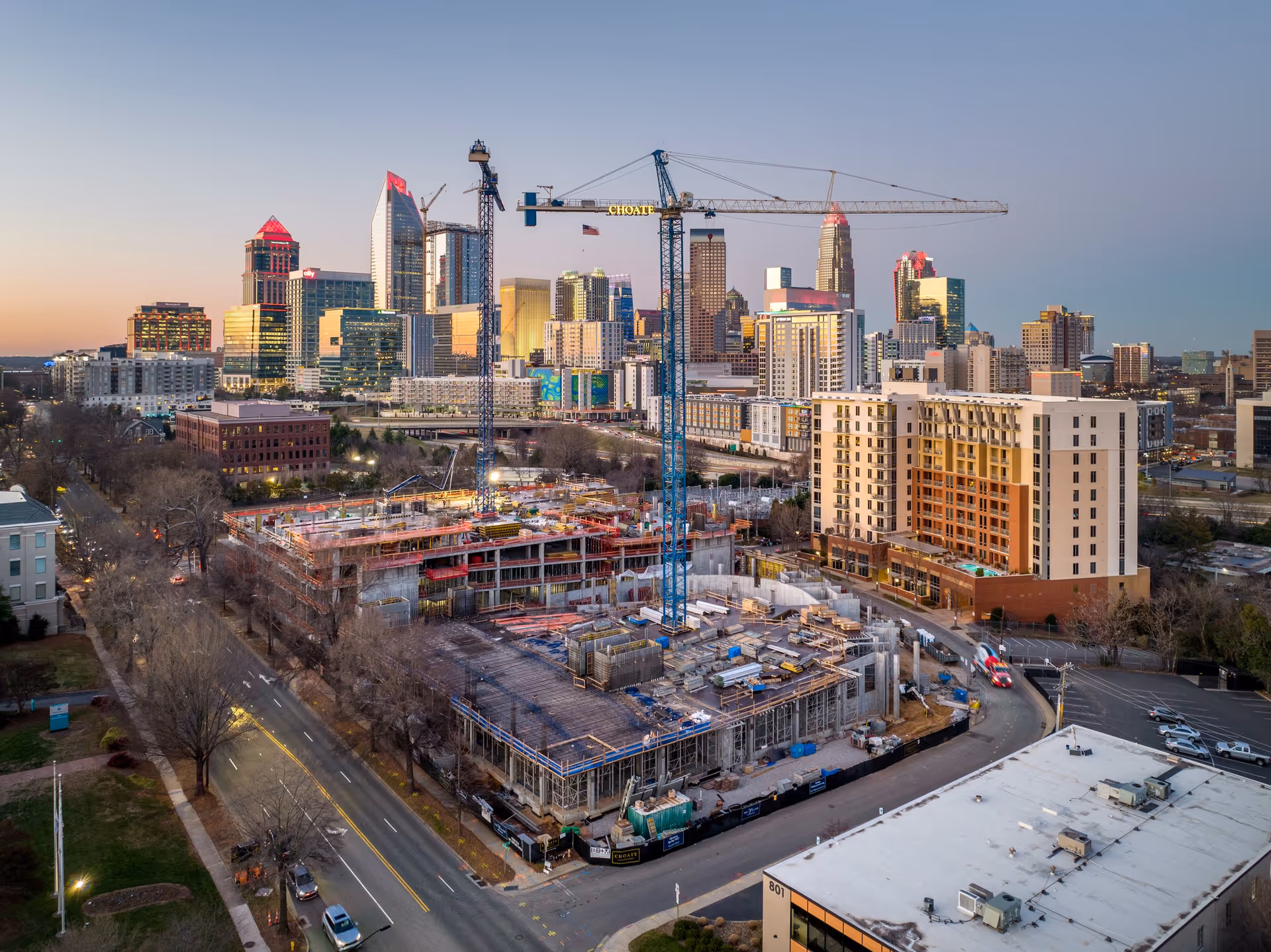 Aerial view of a construction site with cranes in the foreground and a city skyline with tall buildings in the background during sunset.