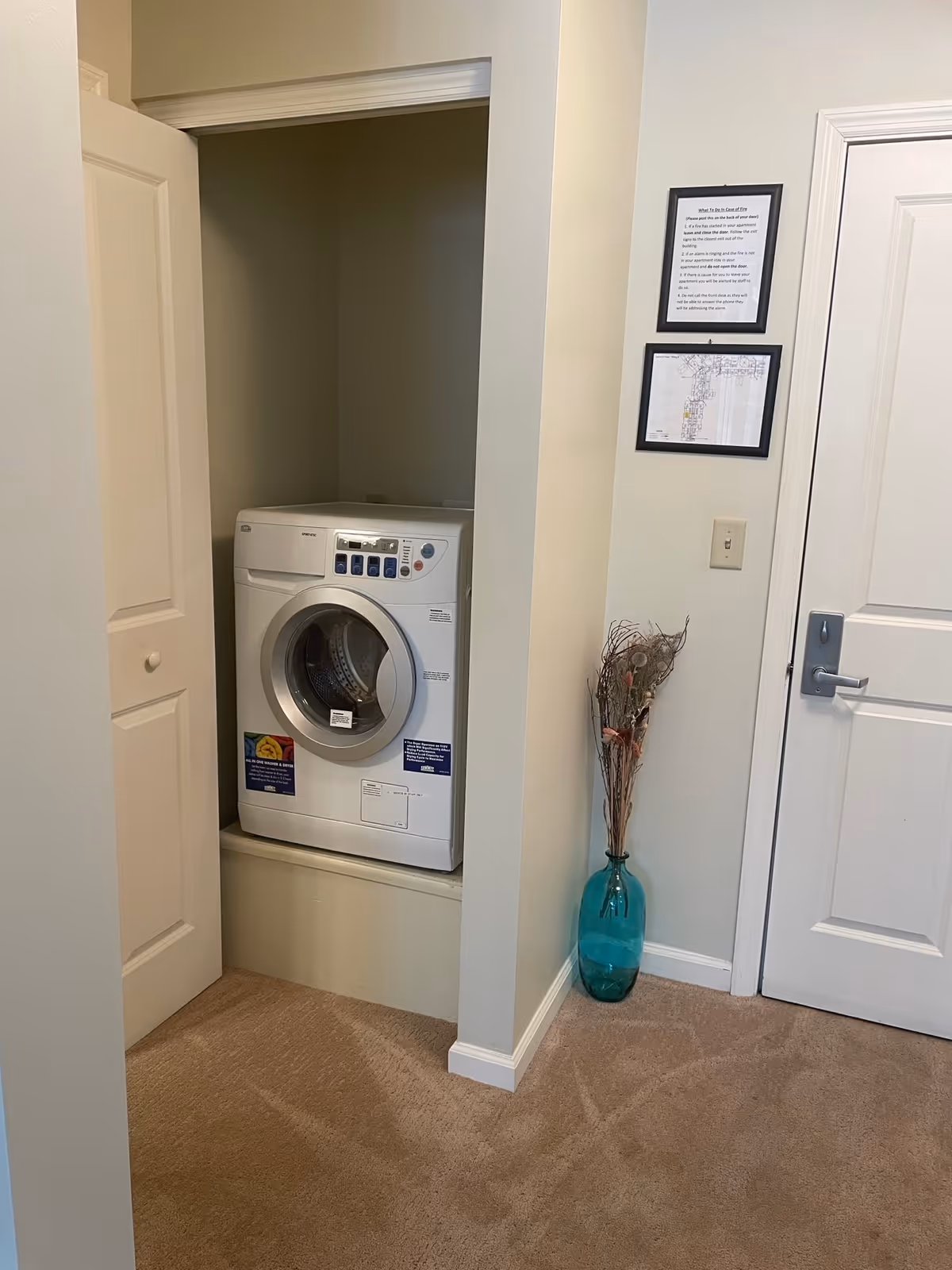 Front-loading washer in a small closet alcove beside a closed door and a teal vase with dried branches.
