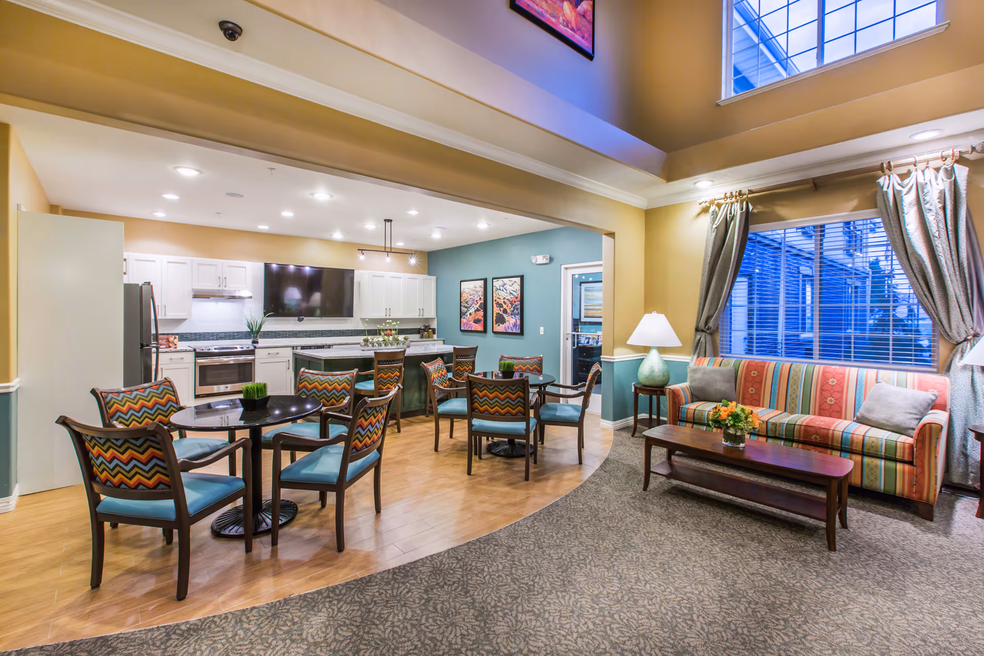 A bright and spacious common area featuring a kitchen and living room. The kitchen has white cabinets, a stainless steel refrigerator, stove, and a large flat-screen TV mounted on the wall. There are multiple round tables with colorful patterned chairs. The living room area includes a striped sofa with cushions, a wooden coffee table with a flower arrangement, two side tables with lamps, and large windows with curtains allowing natural light to enter.