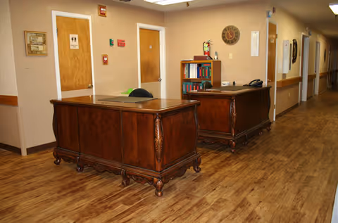 Interior hallway of a nursing-rehab facility with two wooden desks, a chair, a bookshelf with books and a fire extinguisher, a clock on the wall, and two closed wooden doors, one with a restroom sign. The floor is wood-patterned and the walls are beige.