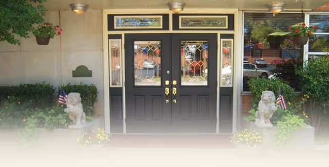 Entrance of a building with double black decorative glass doors flanked by lion statues, potted plants, hanging planters, and small American flags.