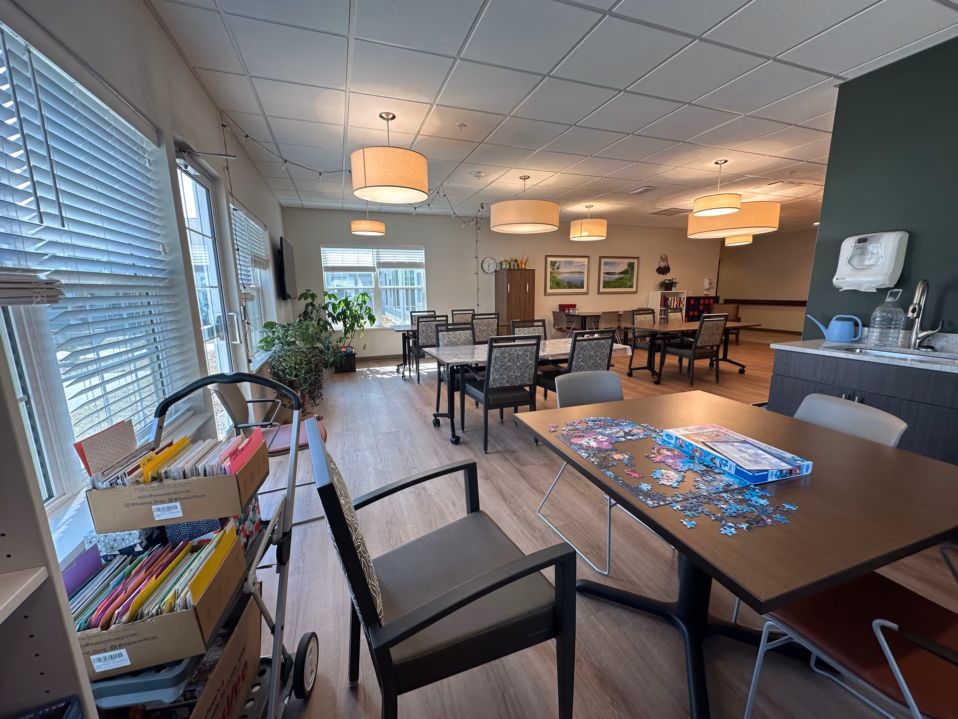 A bright and spacious common area in a senior living community with multiple tables and chairs arranged for group activities. One table has a partially completed jigsaw puzzle and its box. There are large windows with blinds letting in natural light, several hanging ceiling lights, and a cart filled with folders and papers near the windows. The room features wooden flooring, a sink with a water pitcher, and framed nature pictures on the far wall.