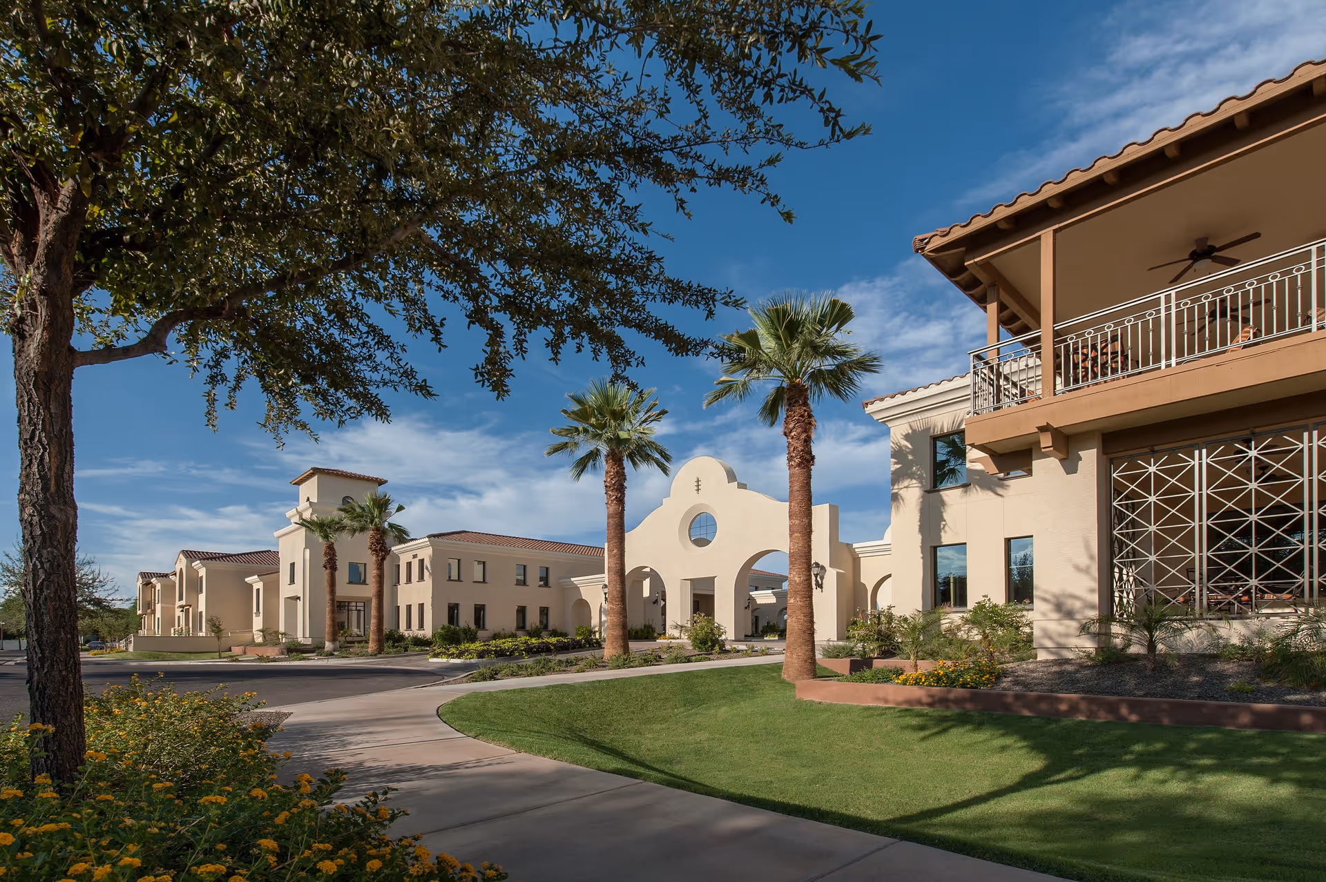 Exterior view of Clearwater Agritopia facility featuring a curved driveway, palm trees, manicured lawns, and beige stucco buildings with tiled roofs under a blue sky with scattered clouds.