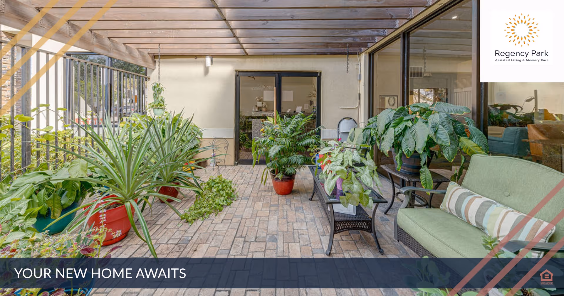 Covered outdoor patio area with brick flooring, various potted plants, a green cushioned bench with a striped pillow, a glass-top table, and a door leading inside the building. The space is enclosed with a metal fence on one side and large windows on the other.