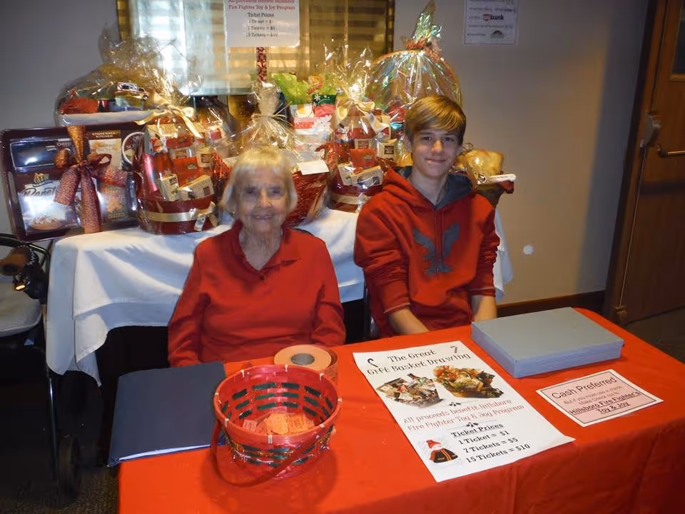 An elderly woman and a teenage boy sitting behind a table covered with a red tablecloth. On the table are raffle tickets, a basket with tickets, and signs for a gift basket drawing fundraiser. Behind them are several gift baskets wrapped in cellophane. The setting appears to be indoors, possibly in a community or senior living facility.