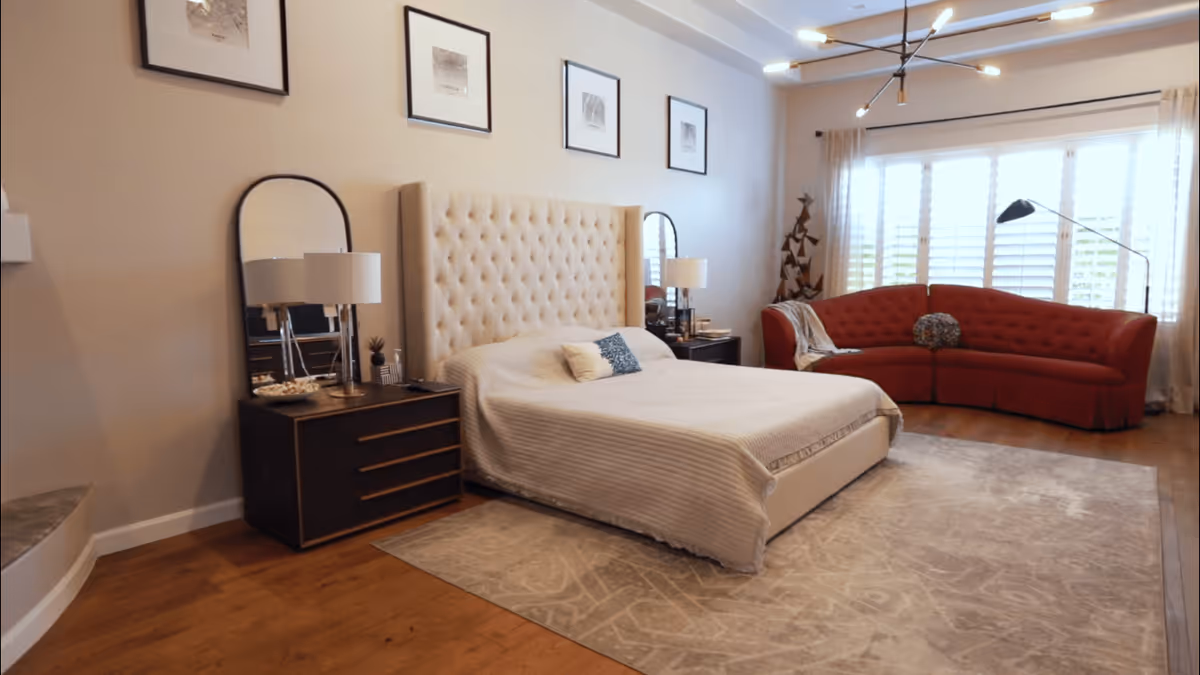 A spacious bedroom with a large tufted beige headboard bed covered with a light-colored bedspread, two dark wooden nightstands with lamps and mirrors behind them, a red curved sofa near a large window with white shutters, and a modern ceiling light fixture.
